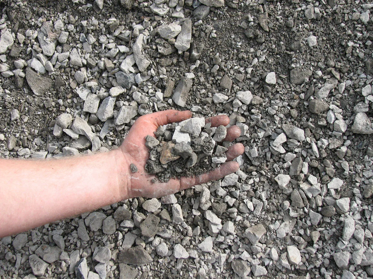A hand holding a scoop of crusher run, angular gray stone mixed with fine stone dust, over a pile of the same material