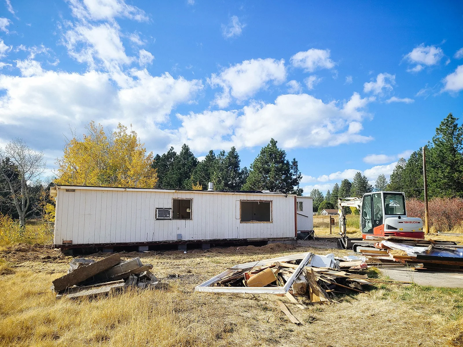 Single-wide mobile home stripped to the shell with the windows out, a mini excavator parked behind it and a pile of pulled framing and trim in front