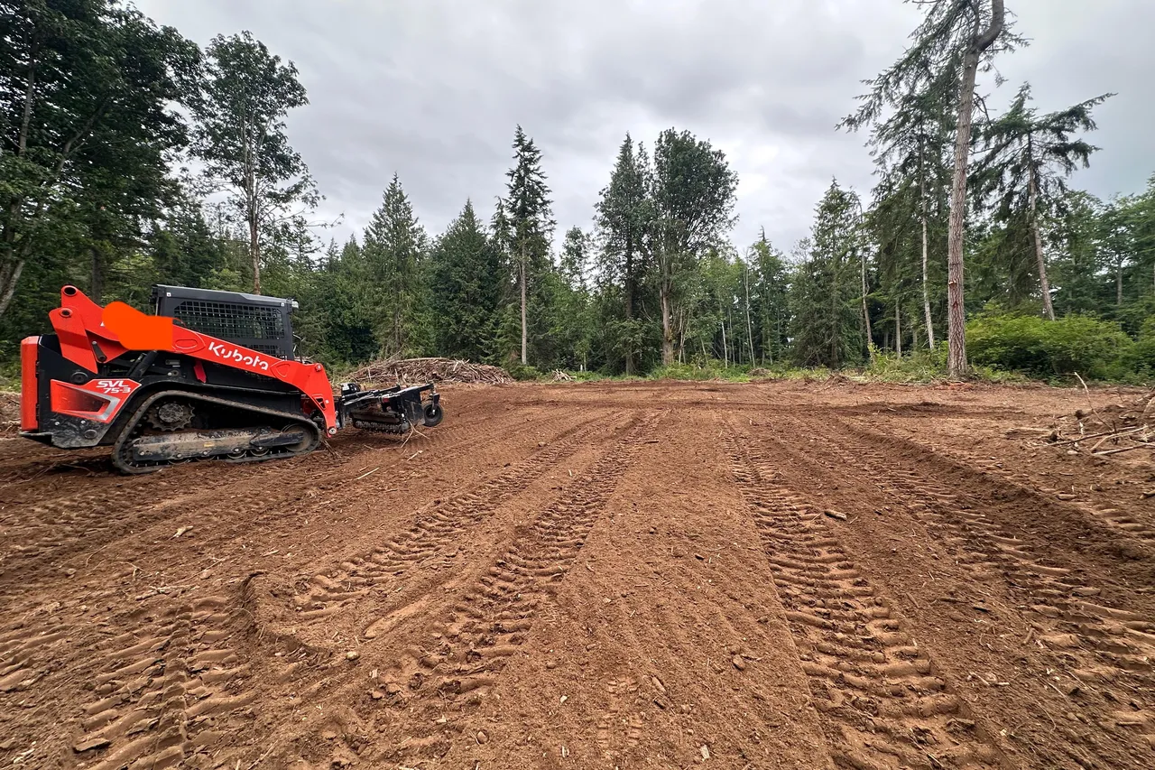 Orange compact track loader on a freshly cleared and graded dirt lot, tread marks across the ground and a tree line behind