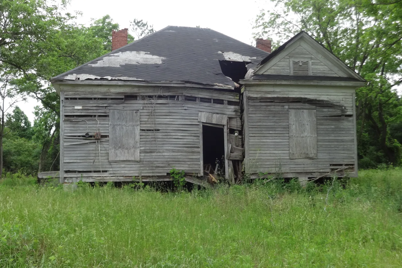 Weathered wood-frame house with a sagging roof, missing shingles and boarded-up windows, standing in tall grass